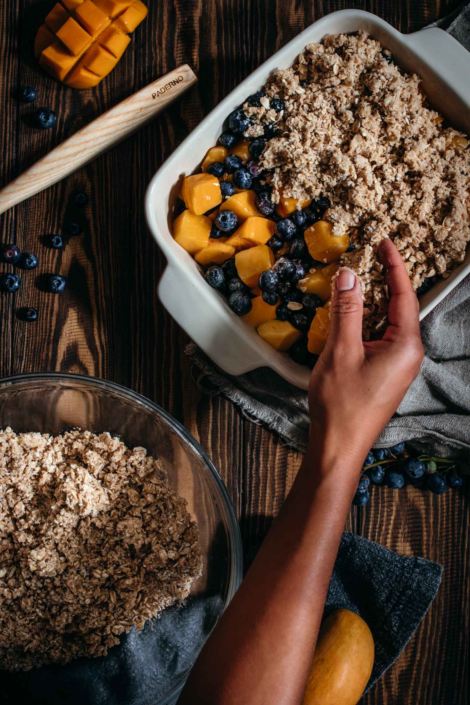 Oat crumble being sprinkled onto blueberries and mangoes