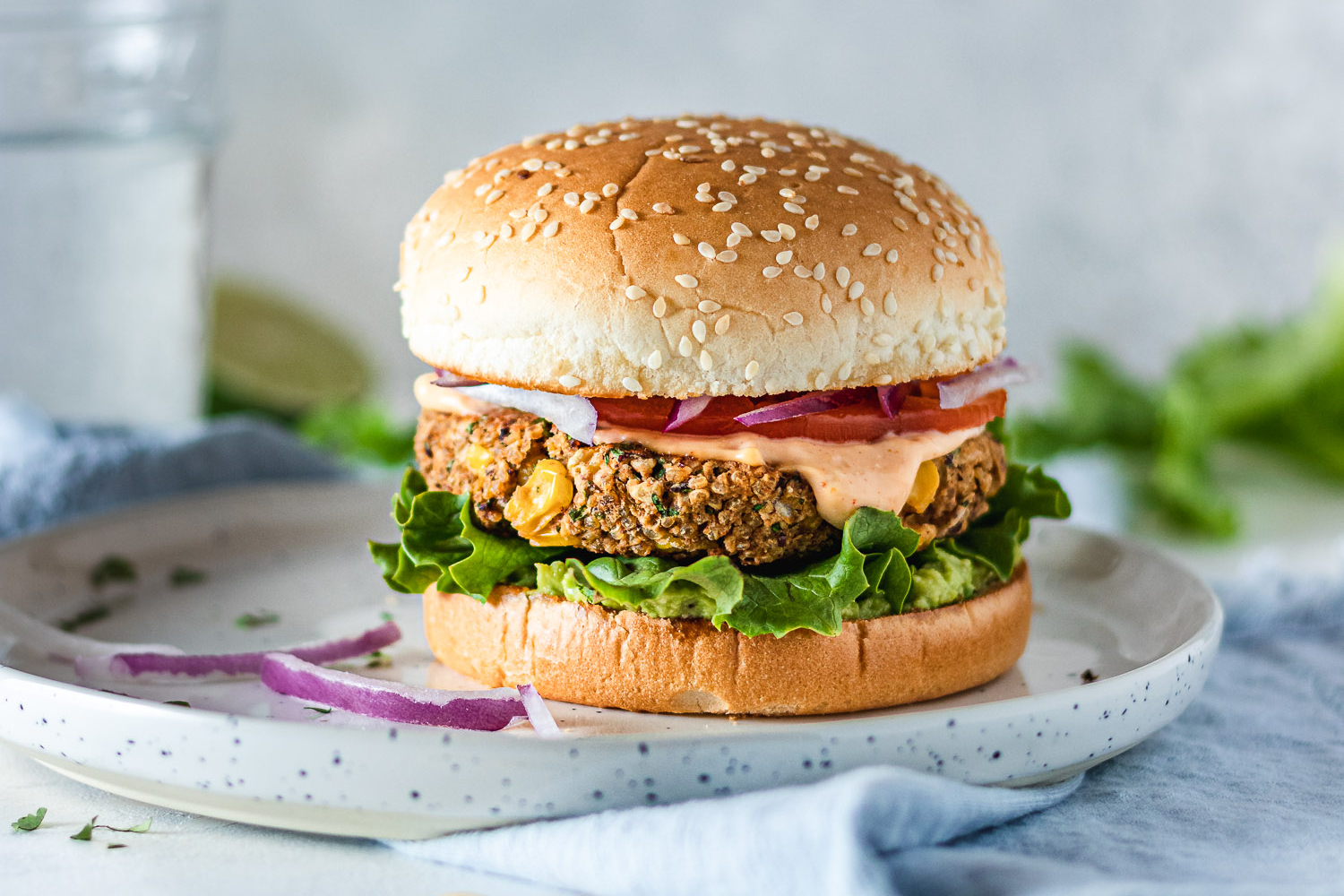 Veggie burger on a plate surrounded by chickpeas, slices of onion, lettuce, a glass of water and half a lime.