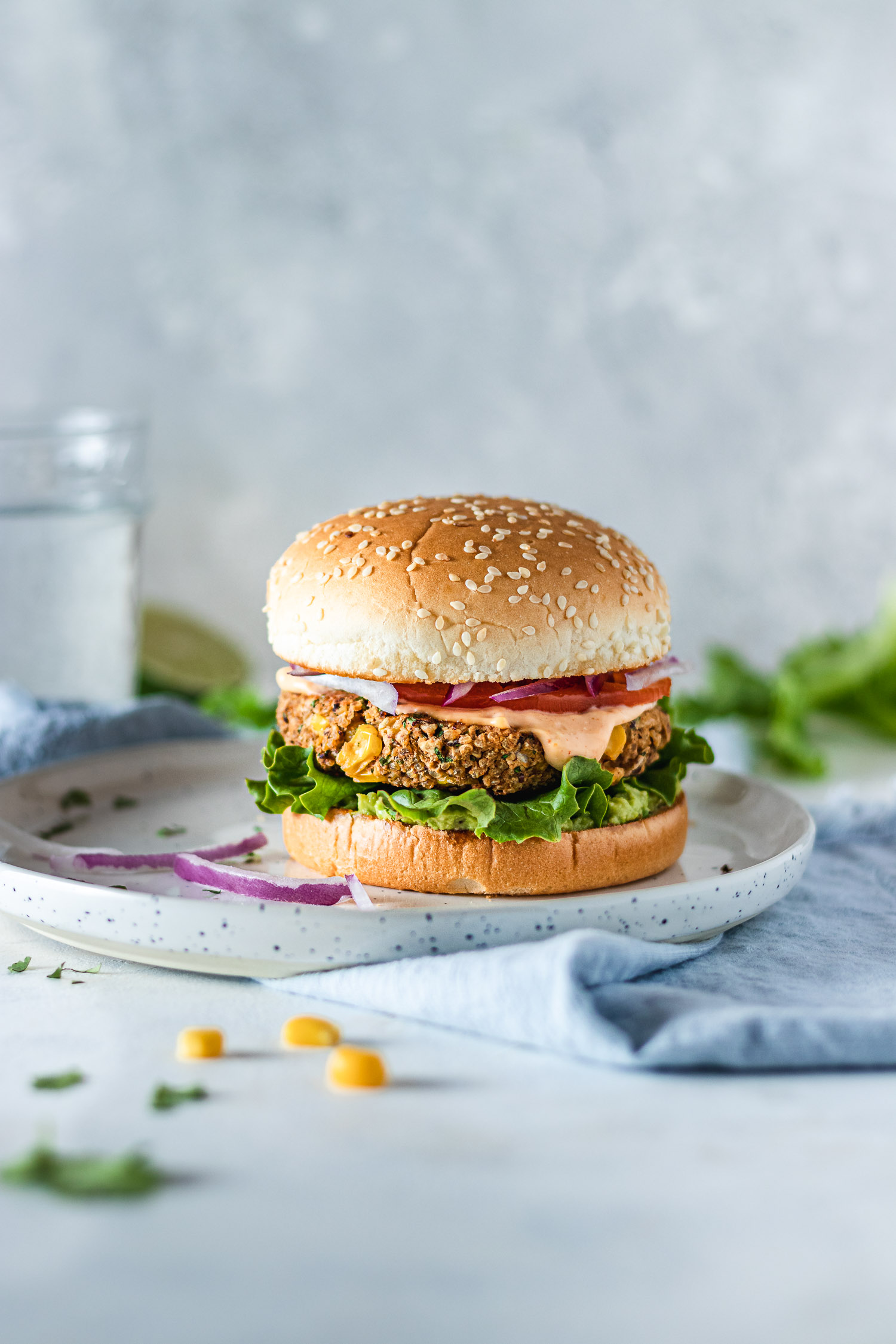 Veggie burger on a plate surrounded by chickpeas, slices of onion, lettuce, a glass of water and half a lime.