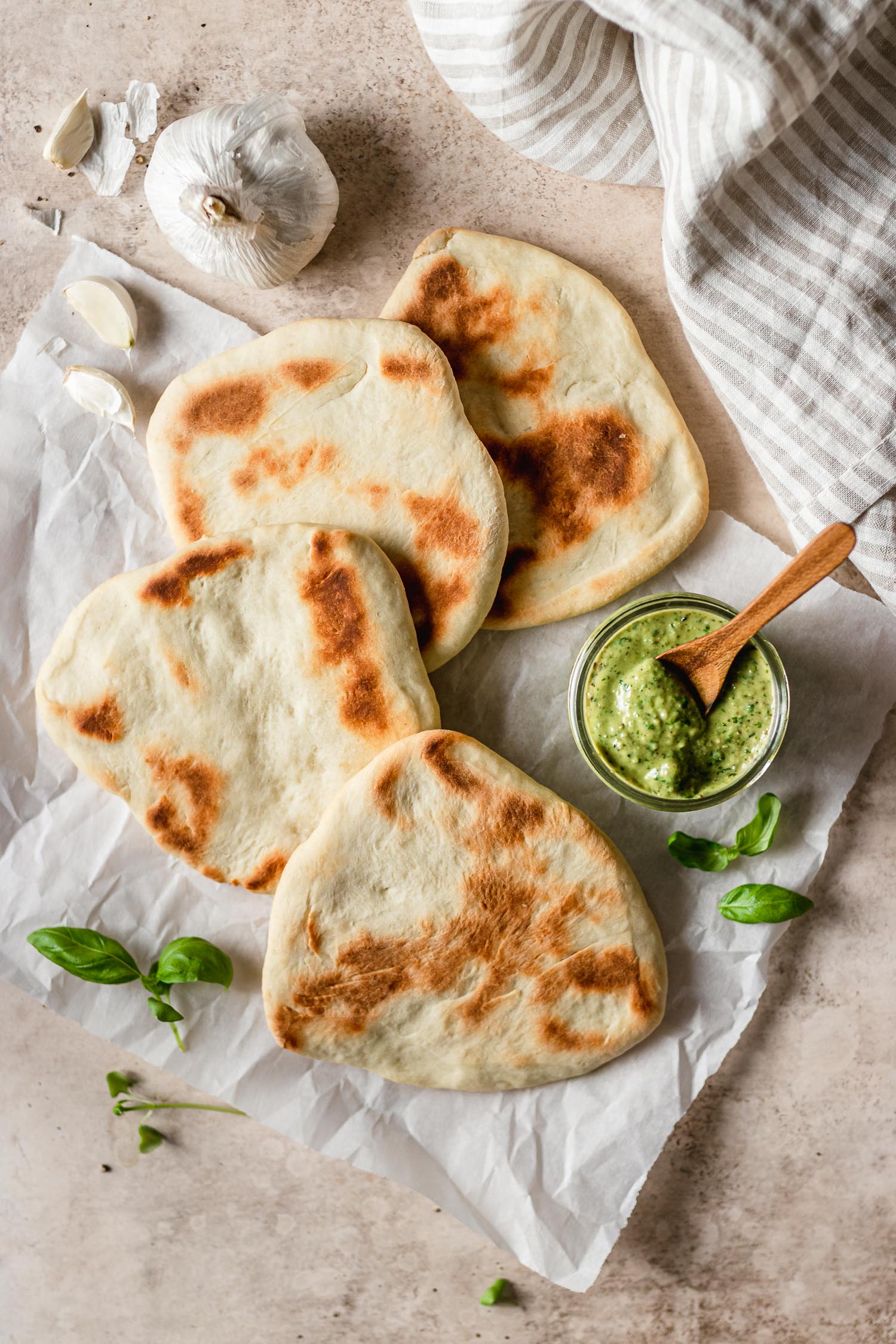 Homemade flatbreads laid flat on surface, surrounded by pesto, basil and fresh garlic