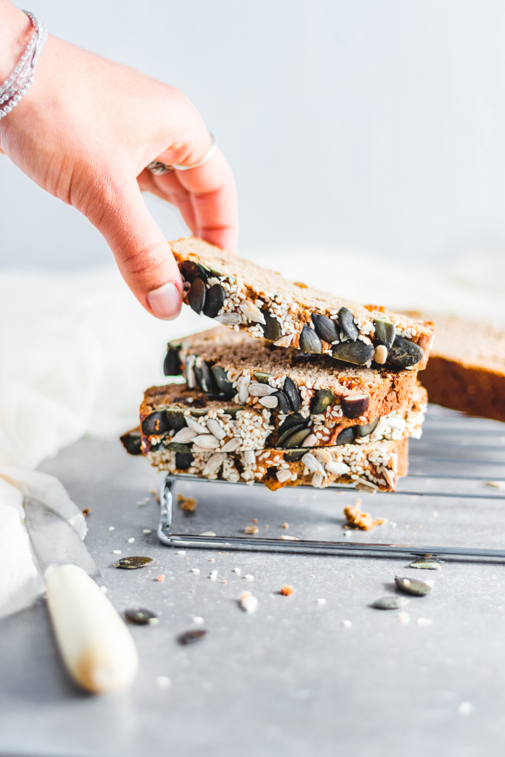 Banana bread slices stacked one on top of the other with sesame seeds, sunflower seeds, and pepitas as garnish. Hand reaching out for a slice