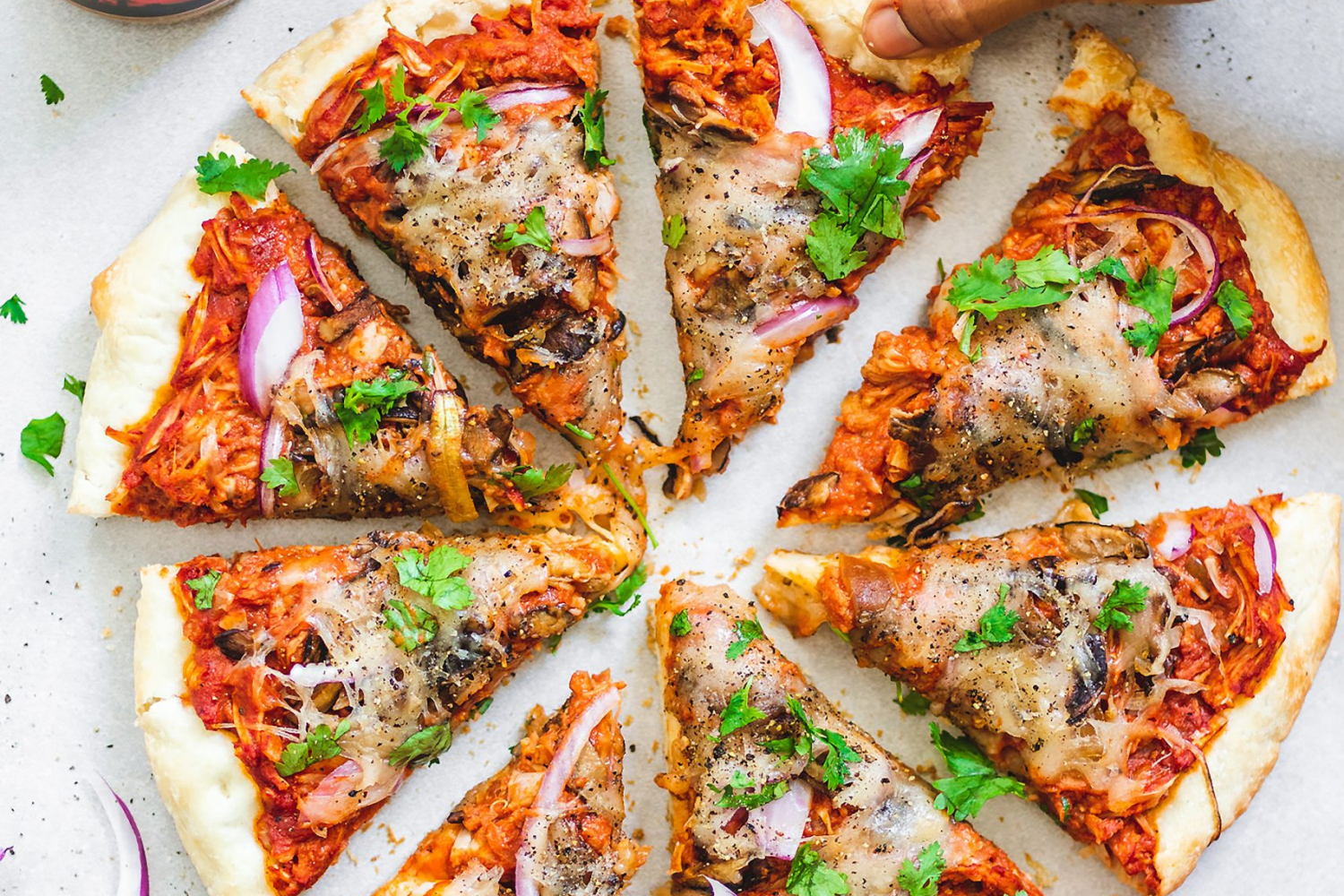 Sliced Peanut Butter and BBQ Jackfruit Pizza surrounded by a jar of peanut butter, tomatoes, red onion, and coriander