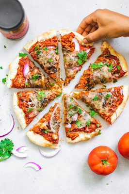 Sliced Peanut Butter and BBQ Jackfruit Pizza surrounded by a jar of peanut butter, tomatoes, red onion, and coriander