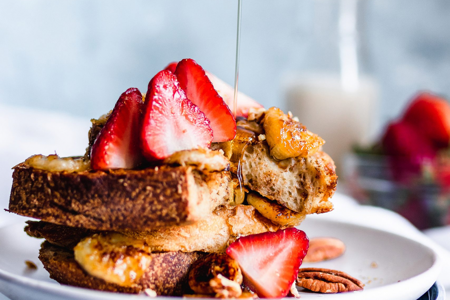 Maple syrup being poured onto a stack of vegan French toast served with caramelized bananas, strawberries, and pecans