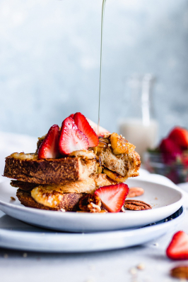 Maple syrup being poured onto a stack of vegan French toast served with caramelized bananas, strawberries, and pecans