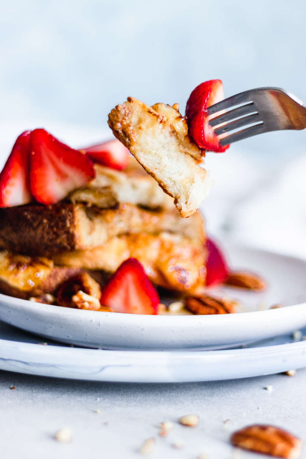 Fork taking a piece of French toast from a stack served with caramelized bananas, strawberries, and pecans
