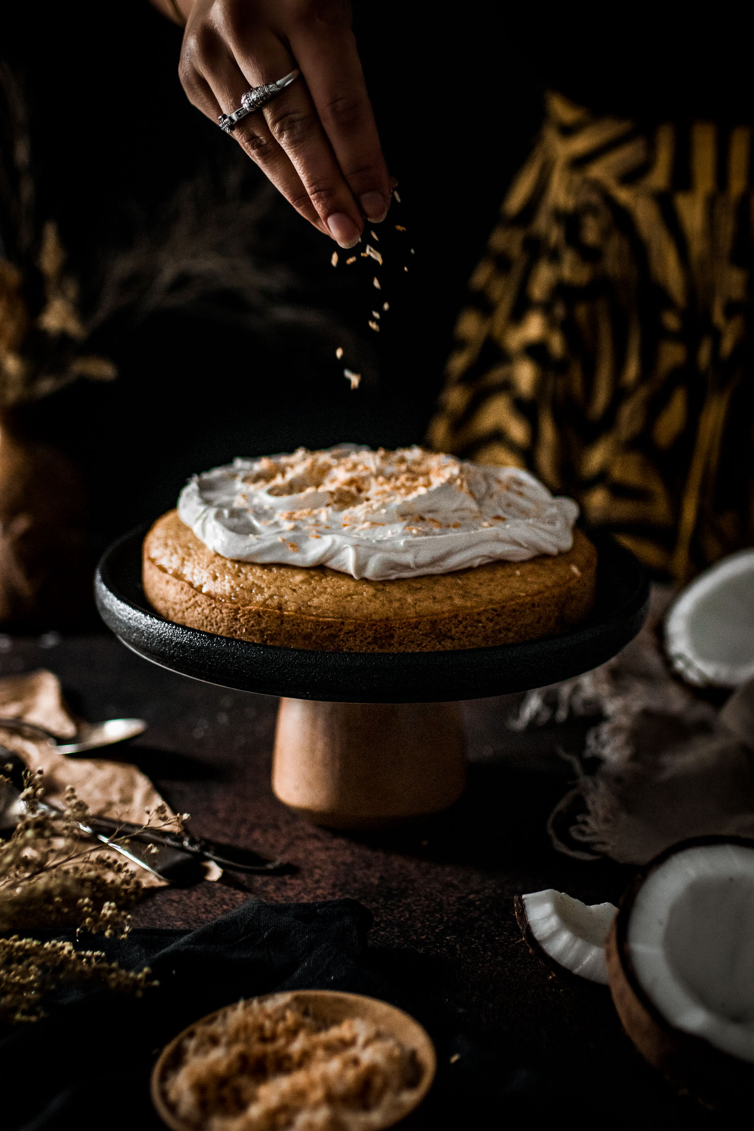 Coconut cake topped with whipped cream being sprinkled with toasted coconut.