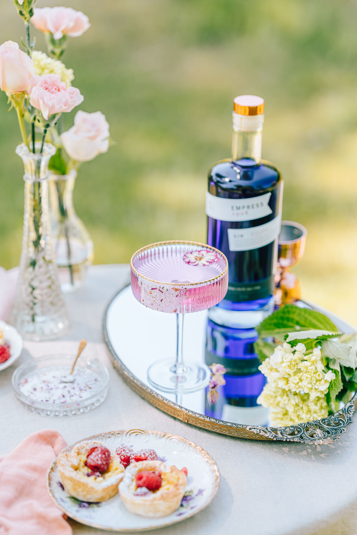 Coupe of rose gin tonic in a garden set on a mirror with bottle of Empress Gin 1908 in the background