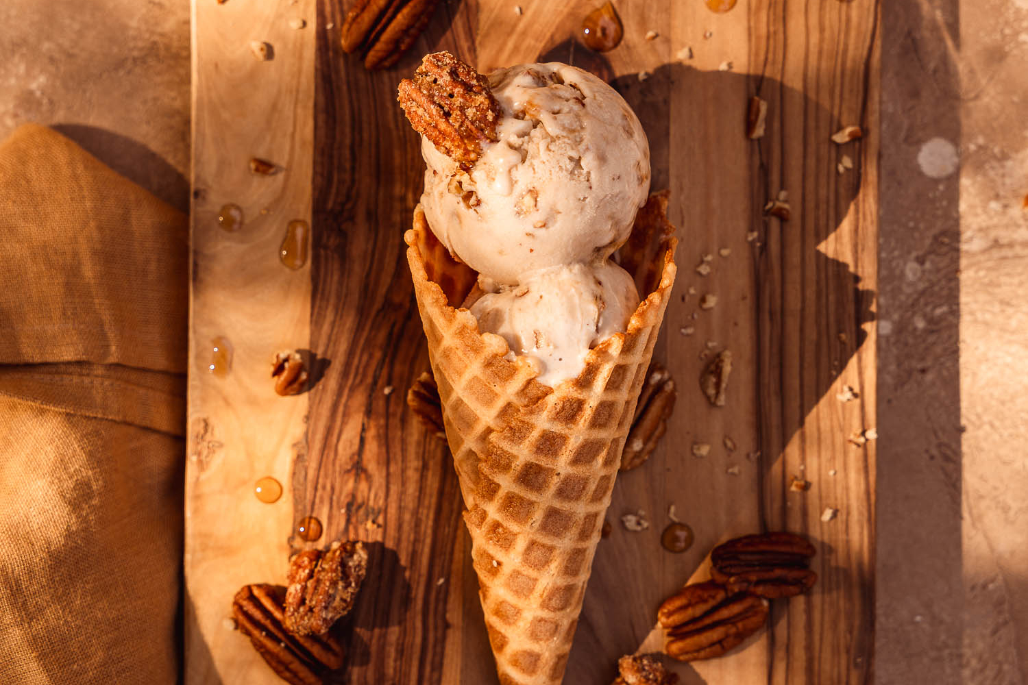 Vegan Maple Pecan Ice Cream cone laying down on a wooden board surrounded by toasted pecans and candied pecans