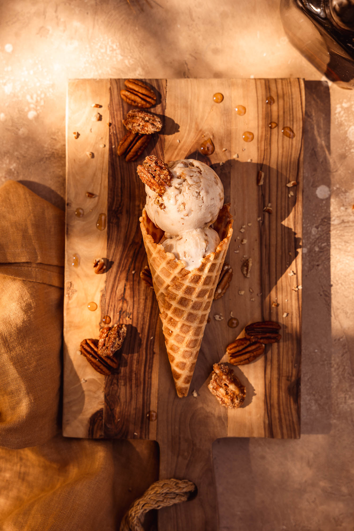 Vegan Maple Pecan Ice Cream cone laying down on a wooden board surrounded by toasted pecans and candied pecans